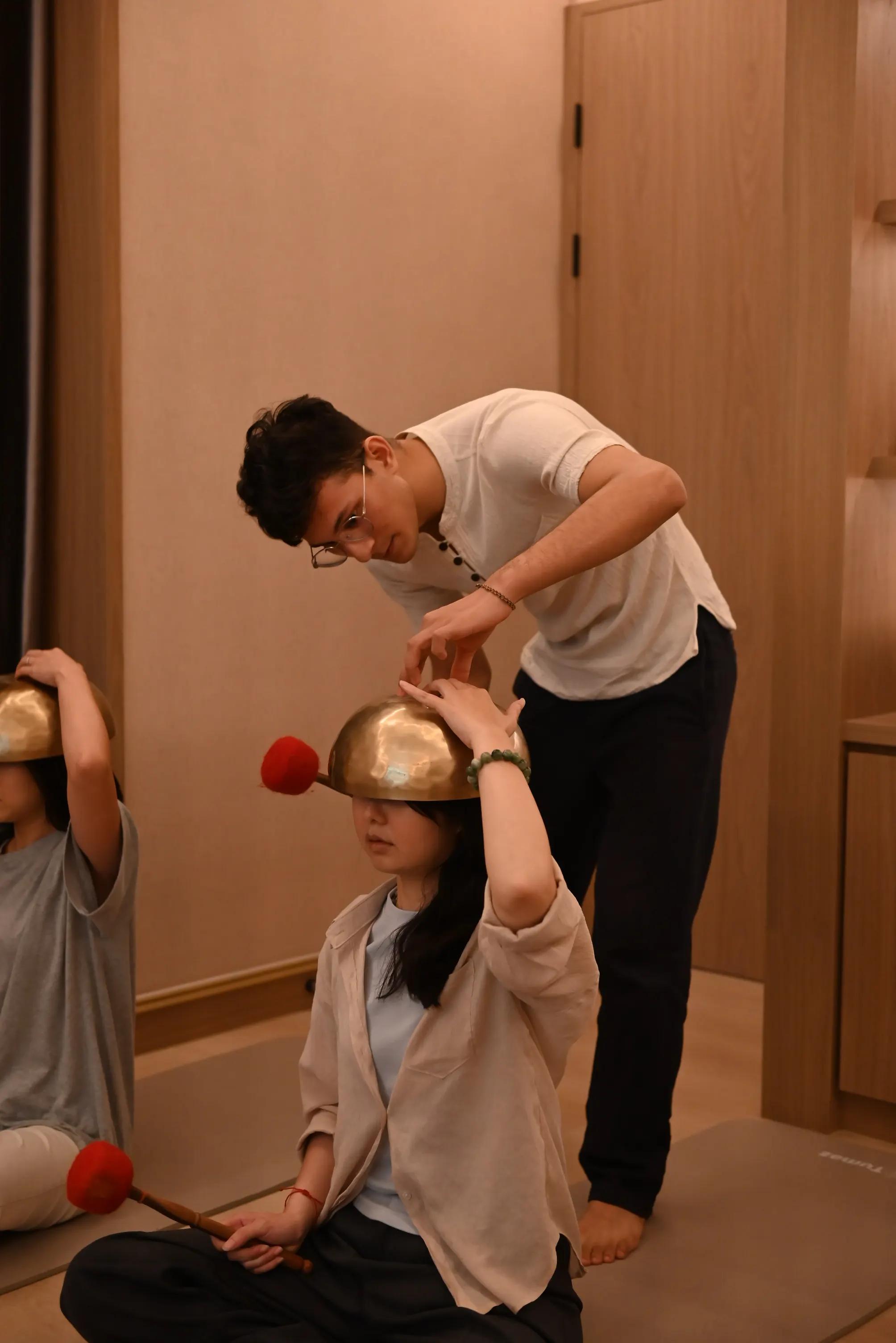 A practitioner in white holds an inverted singing bowl above a seated woman's head while she steadies it with both hands
