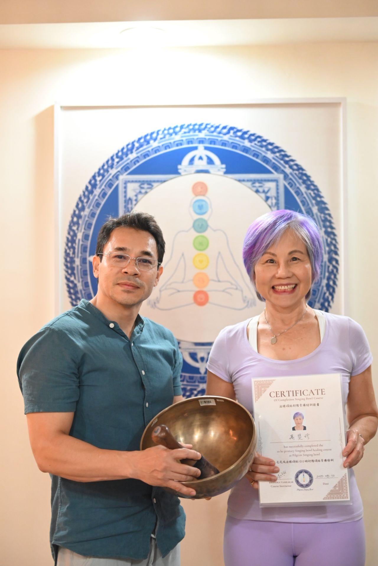 An instructor holding a hand-hammered singing bowl beside a graduate holding her course completion certificate, before a framed chakra mandala